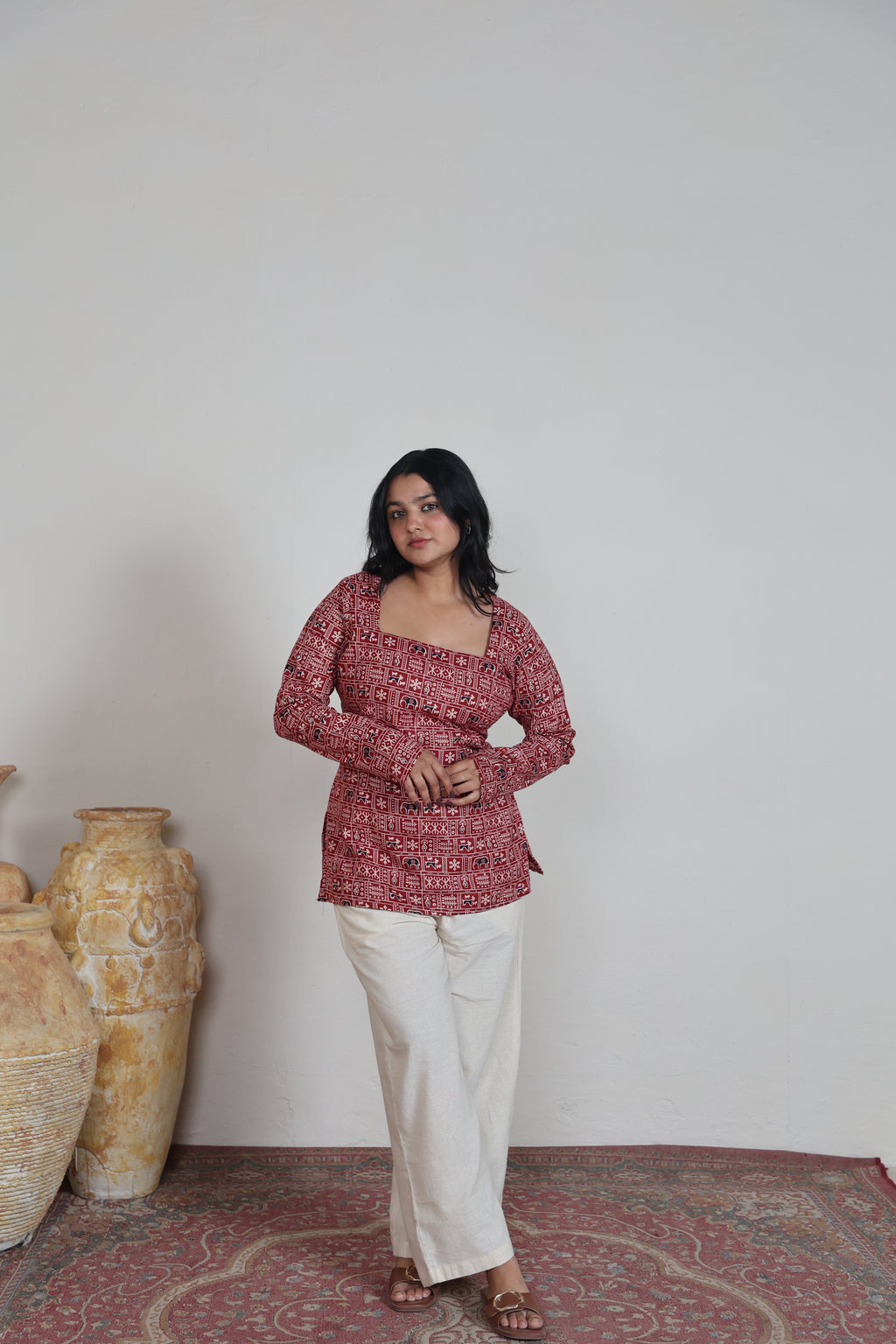 Woman wearing a red patterned top and white pants standing on a patterned rug with vases in the background.