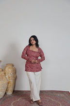 Woman wearing a red patterned top and white pants standing on a patterned rug with vases in the background.