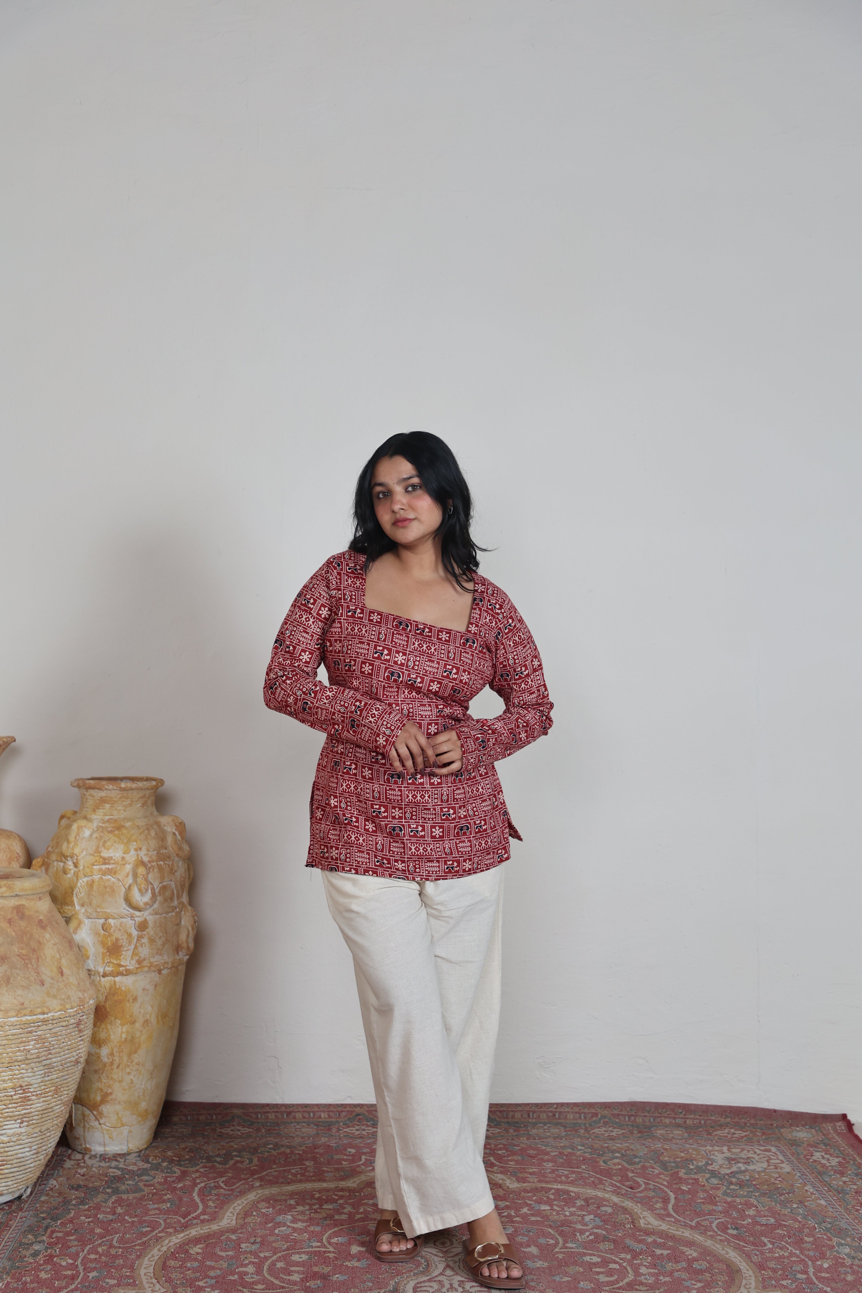 Woman wearing a red patterned top and white pants standing on a patterned rug with vases in the background.