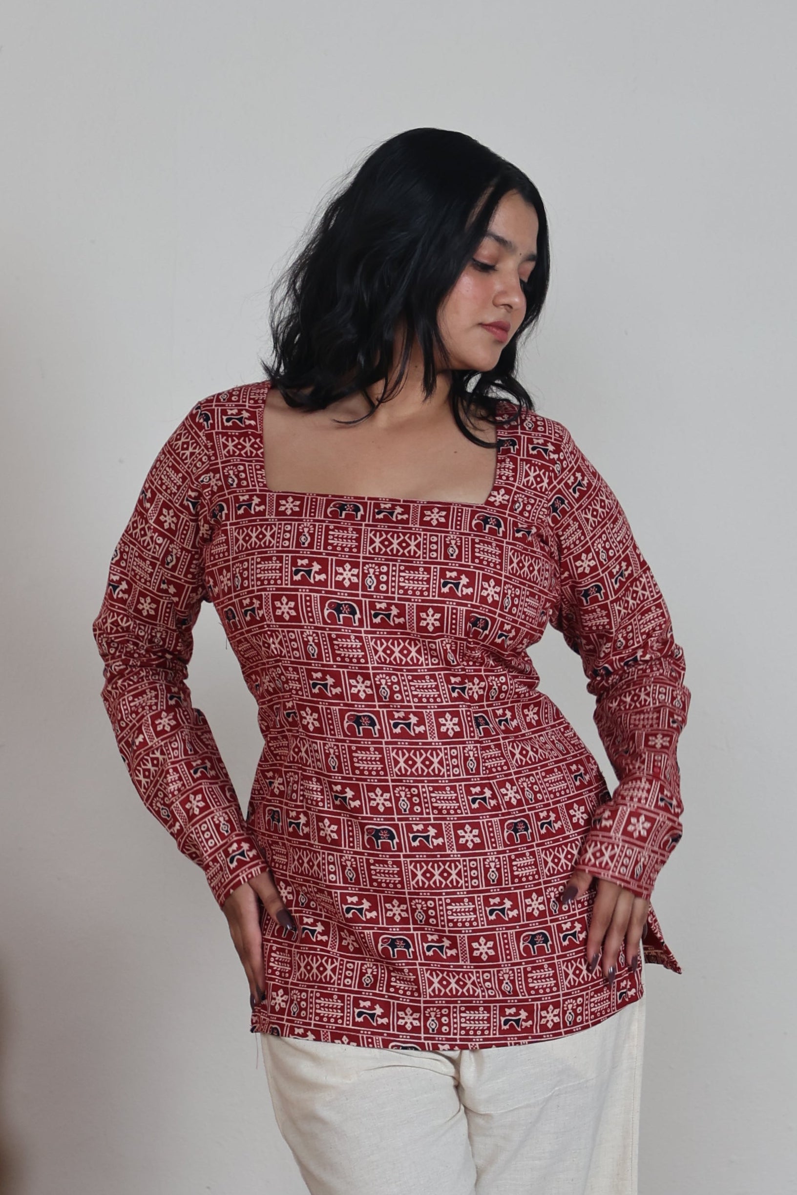 Woman in a red patterned top and white pants standing on a patterned rug with a plain wall background
